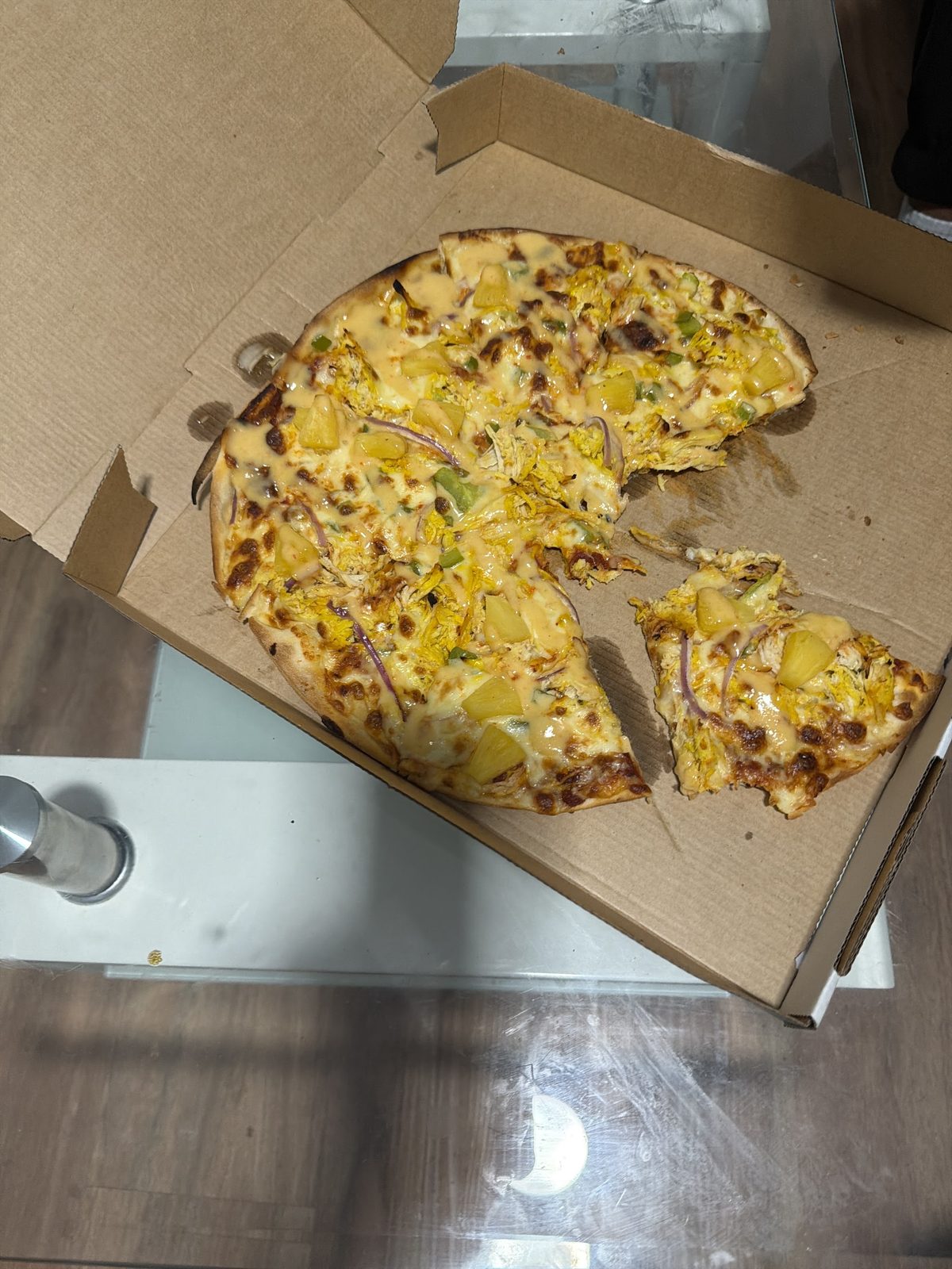 A chicken and pineapple pizza in an open takeaway box, one slice pulled out